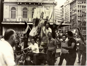 24 Ioυλίου 1974, Αθήνα, Ομόνοια. Πολίτες με σημαίες και μια τεράστια εικόνα του «Μεγάλου Αρχιερέως» πανηγυρίζουν την επιστροφή της δημοκρατίας και του Κωνσταντίνου Καραμανλή.